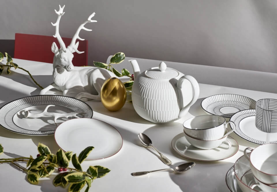 Festlich gedeckter Tisch mit weißer Porzellanfigur eines Hirsches, goldener Ei-Objekt, grafisch gemustertem Service und rankenden grünen Zweigen.
Festive table setting with a white porcelain stag, golden egg object, graphic patterned tableware and trailing green foliage.