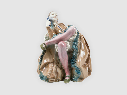 Frontale Ansicht der Porzellanfigur: Dame im Sessel mit übereinandergeschlagenen Beinen, rosa Strümpfen und reich gefälteltem braunem Kleid mit türkisfarbenen Rüschen.
Frontal view of the porcelain figurine: lady in an armchair with crossed legs, pink stockings and a richly pleated brown dress with turquoise ruffles.