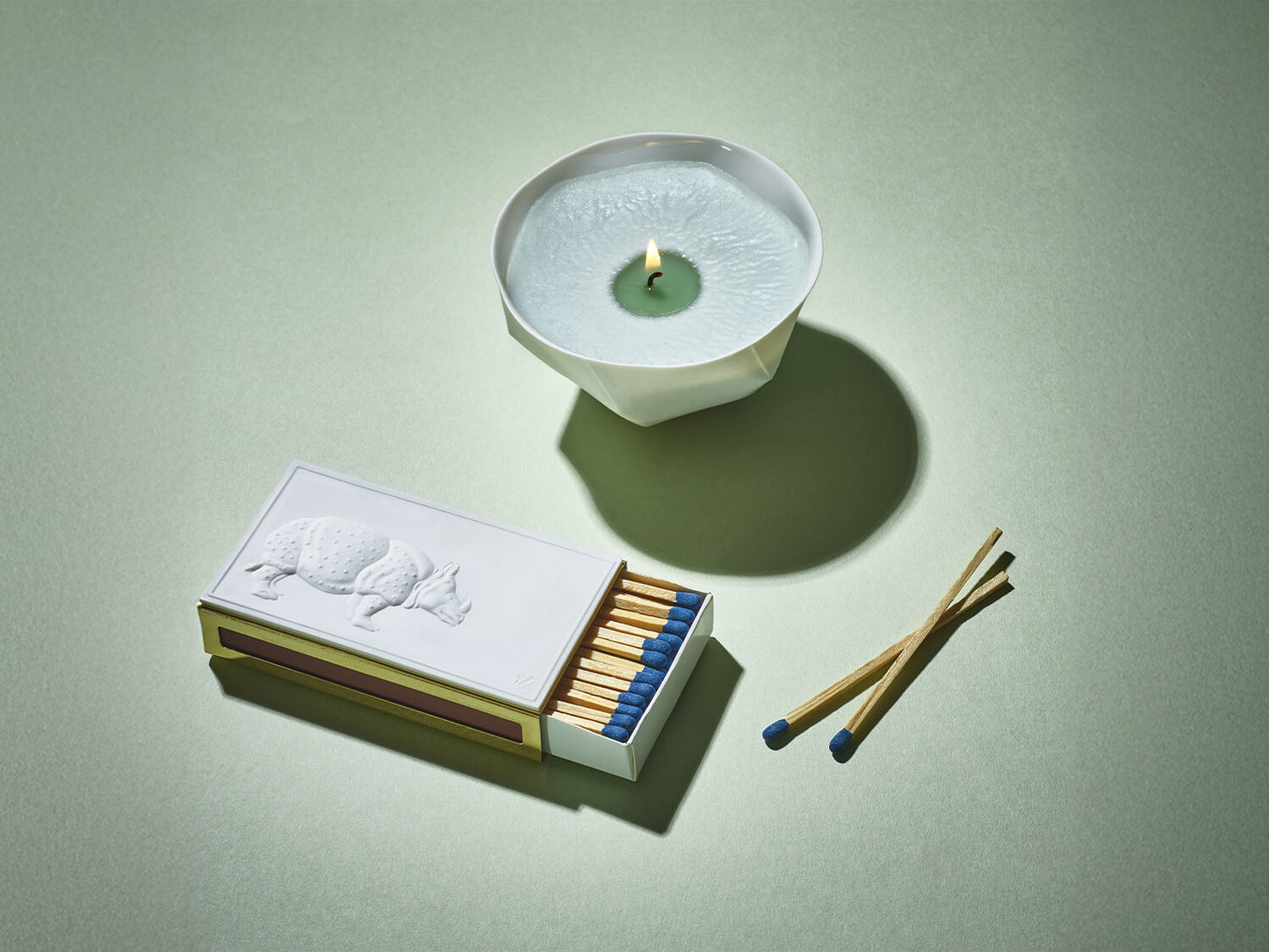 Streichholzetui mit Porzellanrelief und brennender Schale aus weißem Porzellan mit hellgrüner Kerze auf grünem Hintergrund.
Matchbox case with porcelain relief and a burning white porcelain bowl candle on a soft green background.