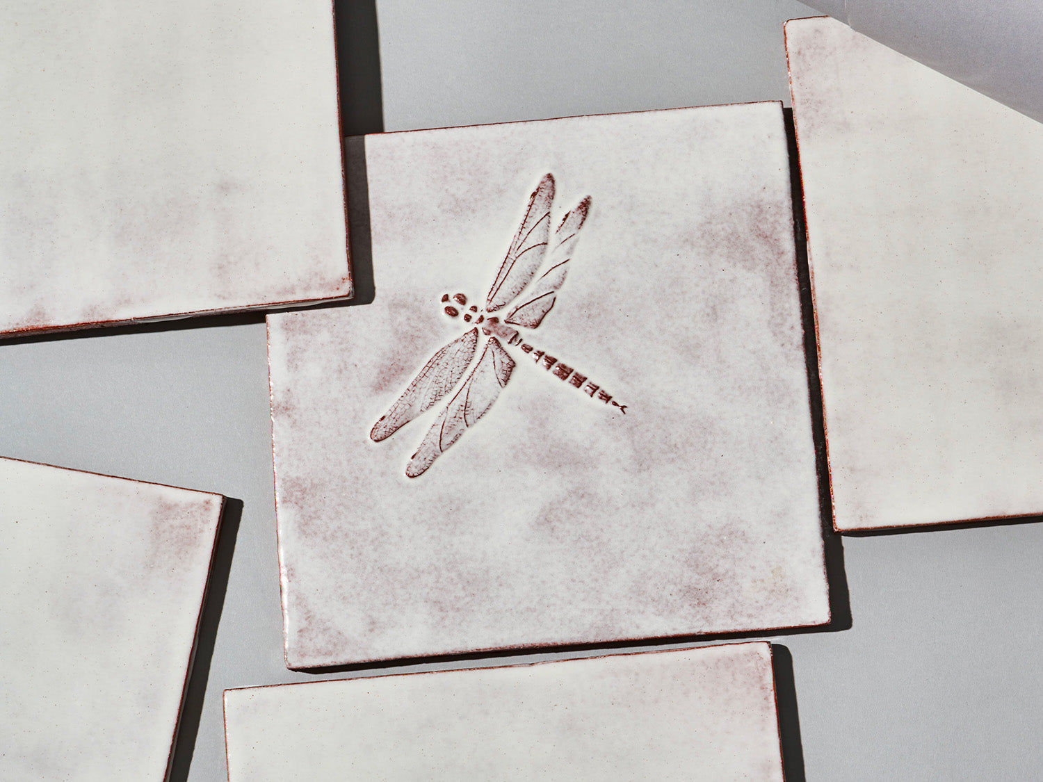 Quadratische, weiße Majolika-Fliese mit leichtem Verlauf und erhabenem Relief einer filigranen Libelle im Zentrum.
Square white-coloured majolica tile with subtle shading and a raised relief of a delicate damselfly in the centre.