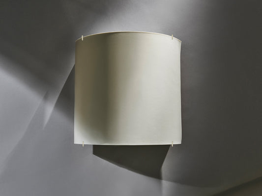 Wandleuchte aus unglasiertem Biskuitporzellan mit schlichter, gewölbter Form vor grauer Wand, unbeleuchtet.
Wall light made of unglazed biscuit porcelain with a simple, curved form mounted on a grey wall, unlit.
