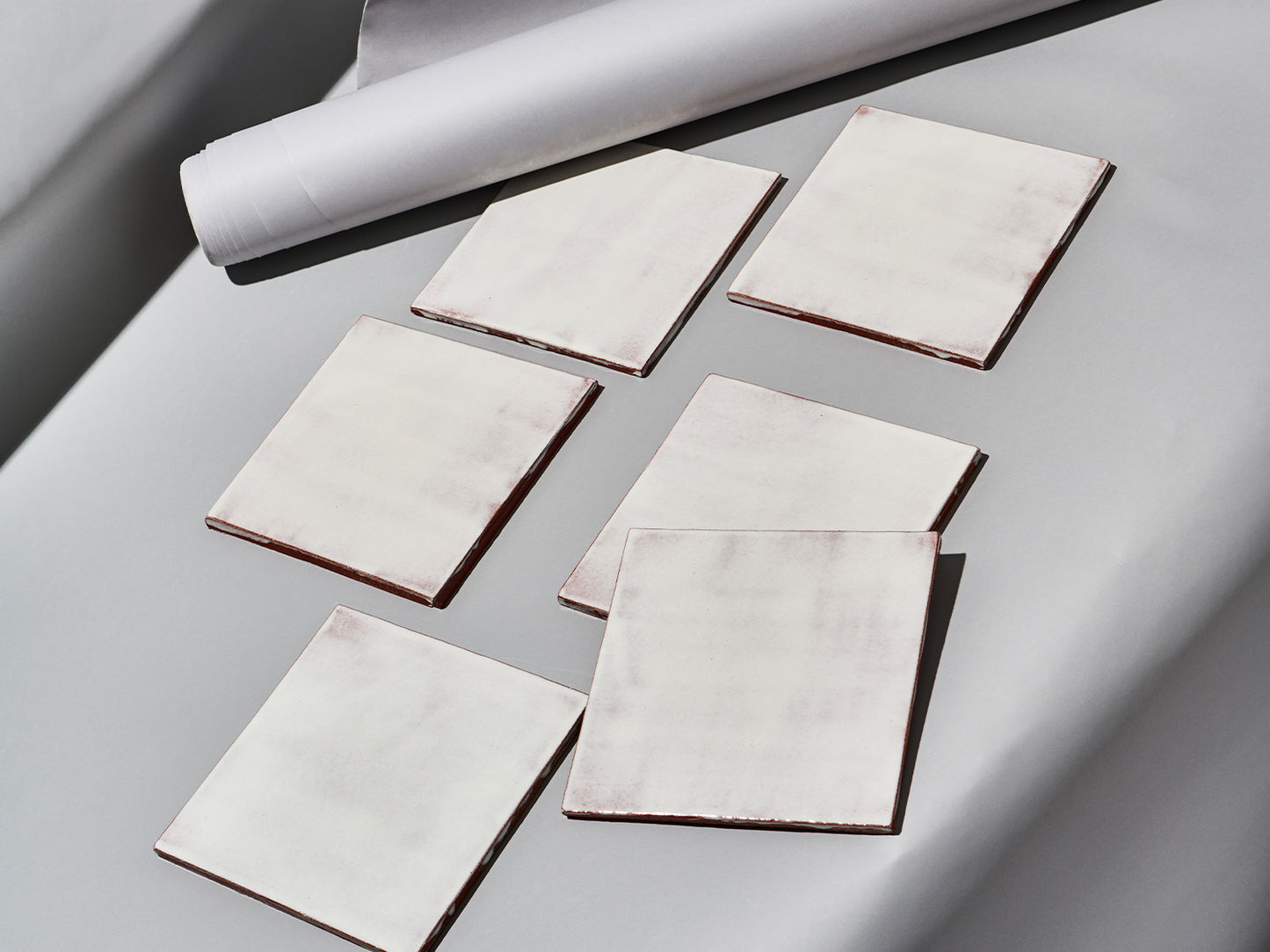 Sechs Majolika-Fliesen mit weißlich-transparenter Glasur „White Frosting“ auf grauem Untergrund arrangiert.
Six majolica tiles with semi-transparent white “White Frosting” glaze arranged on a grey background.