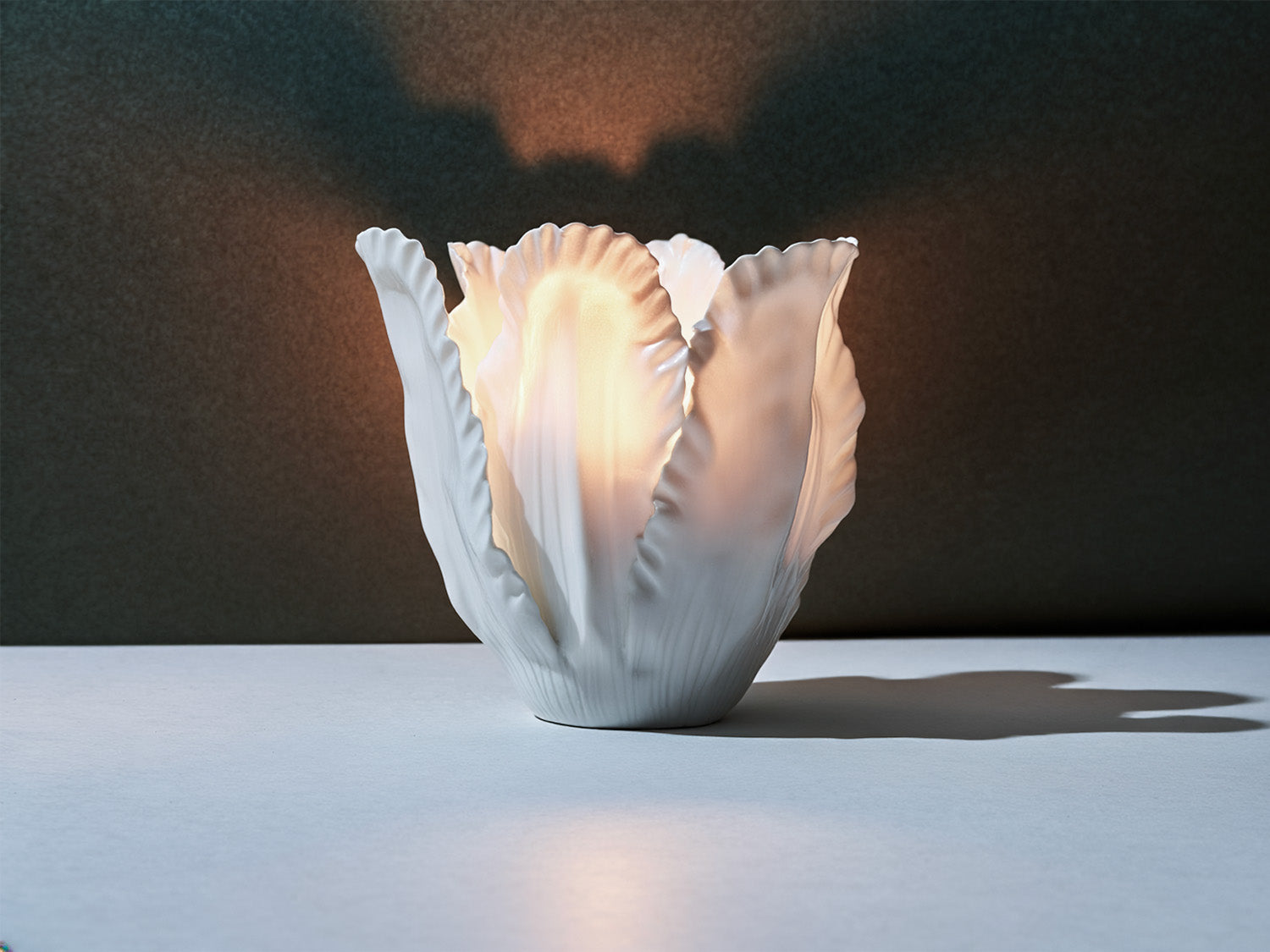 Biskuitporzellan-Windlicht aus überlappenden, blattartigen Segmenten; von innen warm erleuchtet, mit weichem Lichtschein und Schattenwurf.

Bisque-porcelain tea-light with overlapping, leaf-like segments; softly lit from within, casting a warm glow and delicate shadows.