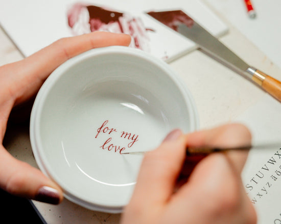 Nahaufnahme: Hände beschriften den Boden einer kleinen weißen Porzellanschale mit roter Kalligraphie „for my love“; feiner Pinsel, Alphabet-/Schriftvorlage und Werkzeuge auf dem Arbeitstisch.


Close-up: hands lettering the words “for my love” in red calligraphy inside a small white porcelain bowl; fine brush, lettering template, and tools on a work table.