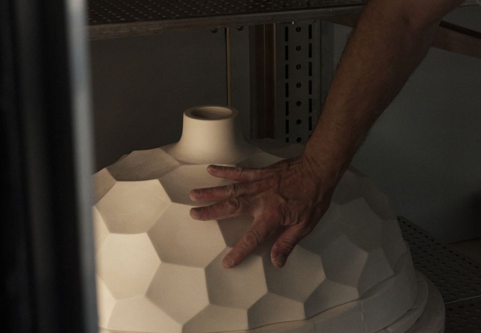 Hand eines Handwerkers stützt einen großen, unglasierten Porzellanschirm mit facettierter Wabenstruktur in einem Regal der Werkstatt.


A craftsman’s hand supports a large, unglazed porcelain shade with a faceted honeycomb structure on a workshop shelf.