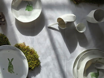 Draufsicht auf eine helle Tischinszenierung mit weißen Porzellangefäßen, Tellern mit grünem Hasenmotiv und Moospolstern.

Top view of a light table arrangement with white porcelain vessels, plates featuring a green hare motif, and patches of moss.