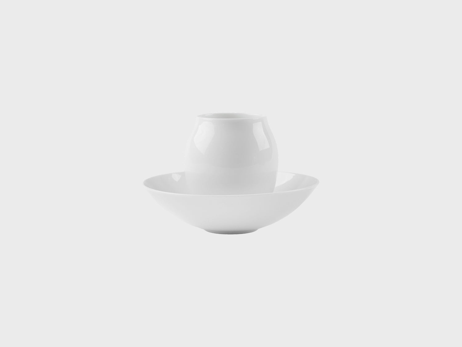 Zwei weiße Porzellanobjekte auf hellem Hintergrund: eine kleiner, bauchiger Mokka Becher steht mittig in einer flachen Schale. Beide Objekte sind schlicht, glänzend glasiert und ohne Dekor.

Two white porcelain objects on a light background: a small, rounded mocca beaker stands centered inside a shallow bowl. Both pieces are simple, glossy glazed, and undecorated.