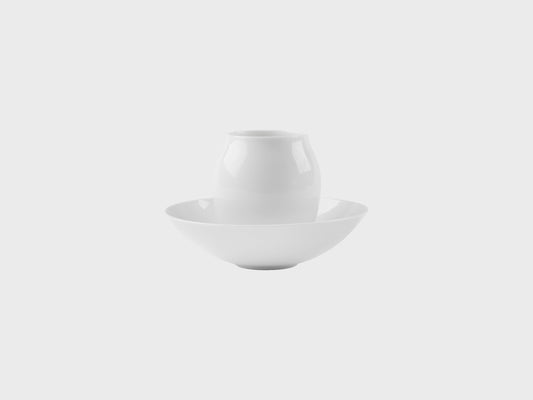 Zwei weiße Porzellanobjekte auf hellem Hintergrund: eine kleiner, bauchiger Mokka Becher steht mittig in einer flachen Schale. Beide Objekte sind schlicht, glänzend glasiert und ohne Dekor.

Two white porcelain objects on a light background: a small, rounded mocca beaker stands centered inside a shallow bowl. Both pieces are simple, glossy glazed, and undecorated.