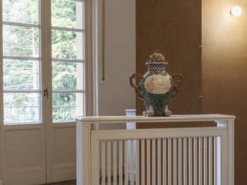 Fertiggestelltes keramisches Gefäß der Künstlerin Nigin Beck auf einem Sockel in einem Ausstellungsraum. Das Werk ist mit organischen Formen, farbigen Glasuren und figürlichen Motiven verziert.

A finished ceramic vessel by artist Nigin Beck displayed on a pedestal in an exhibition space. The work features organic forms, colorful glazes, and figurative motifs.