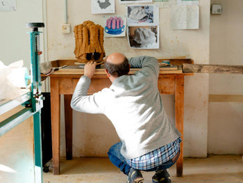 Der Künstler Justin Lieberman kniet vor einem Arbeitstisch in seinem Atelier und bearbeitet ein Tonmodell für ein Kunstwerk der Manifesta 16 Ruhr. Skizzen und Referenzbilder hängen an der Wand hinter dem Modell.

Artist Justin Lieberman kneels at a workbench in his studio while working on a clay model for an artwork created for Manifesta 16 Ruhr. Sketches and reference images are displayed on the wall behind the model.