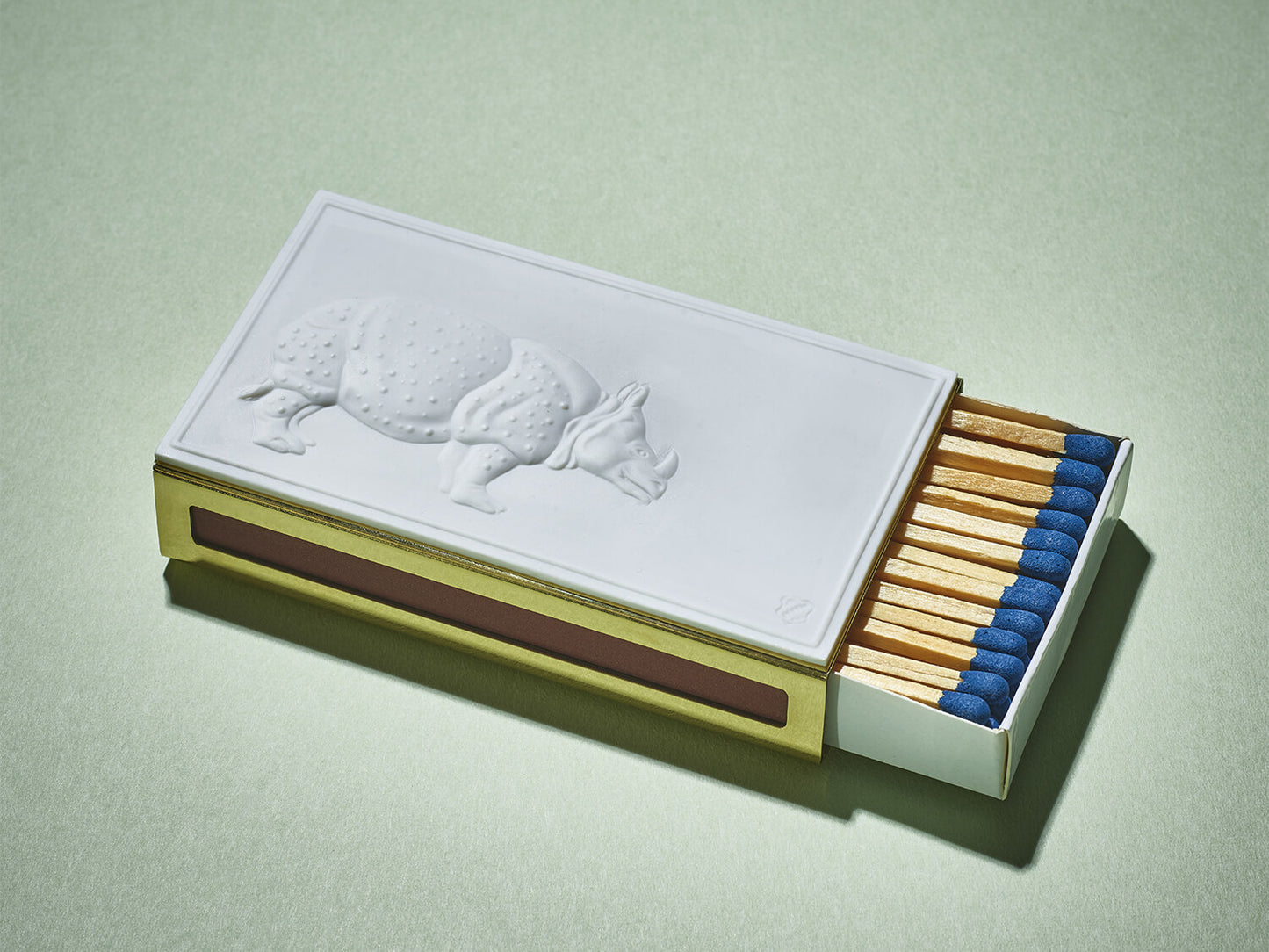 Rechteckiges Streichholzetui mit weißem Porzellanrelief eines Nashorns auf messingfarbener Hülle, blaue Zündholzköpfe sichtbar.

Rectangular matchbox case with white porcelain rhino relief on a brass-coloured sleeve, blue-tipped matches visible.