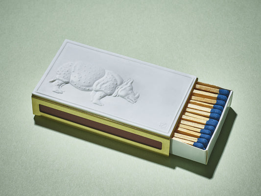 Rechteckiges Streichholzetui mit weißem Porzellanrelief eines Nashorns auf messingfarbener Hülle, blaue Zündholzköpfe sichtbar.

Rectangular matchbox case with white porcelain rhino relief on a brass-coloured sleeve, blue-tipped matches visible.