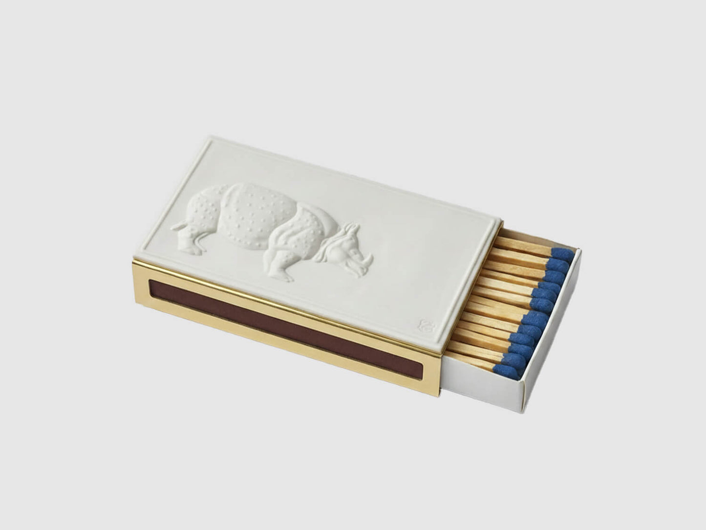 Streichholzetui mit weißer Porzellanplatte und Nashornrelief auf messingfarbener Hülle, Streichhölzer mit blauen Köpfen vor hellem Hintergrund.

Matchbox case with white porcelain plate and rhino relief on a brass-coloured sleeve, blue-tipped matches against a light background.