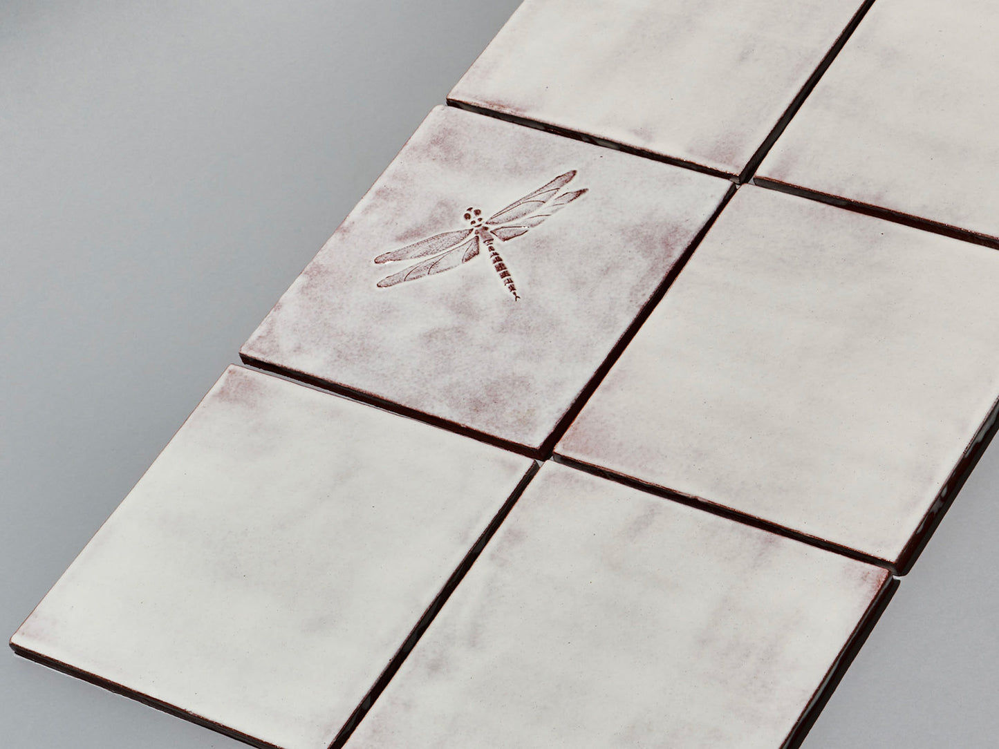 Quadratische, weiße Majolika-Fliese mit leichtem Verlauf und erhabenem Relief einer filigranen Libelle im Zentrum.

Square white-coloured majolica tile with subtle shading and a raised relief of a delicate damselfly in the centre.
